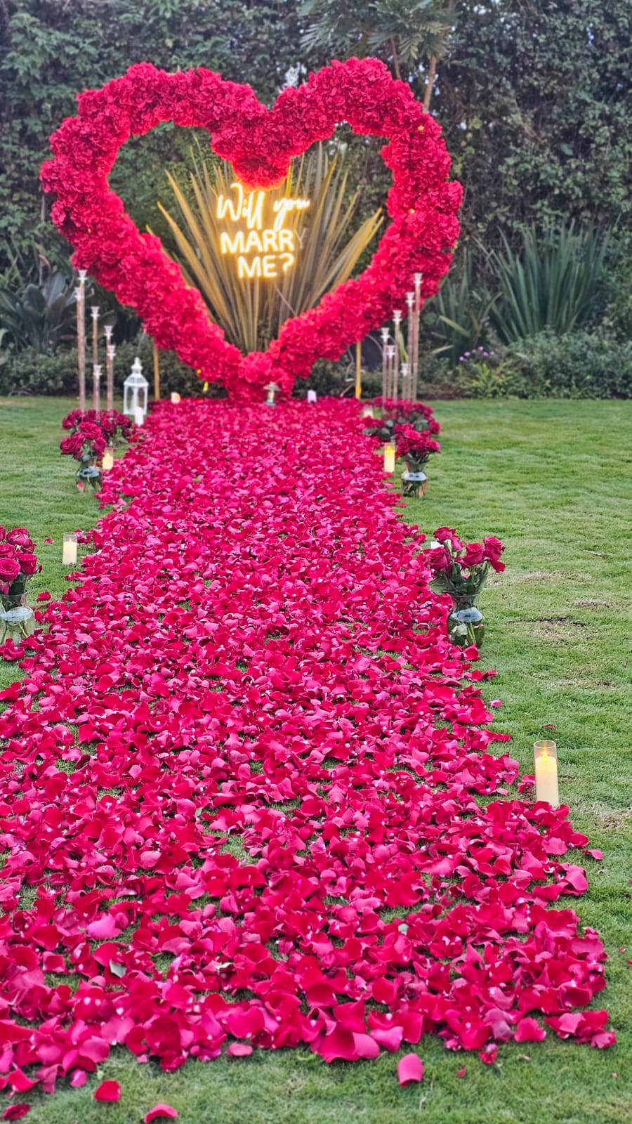 Red rose heart arch with a Will You Marry Me neon sign above a petal-covered garden walkway