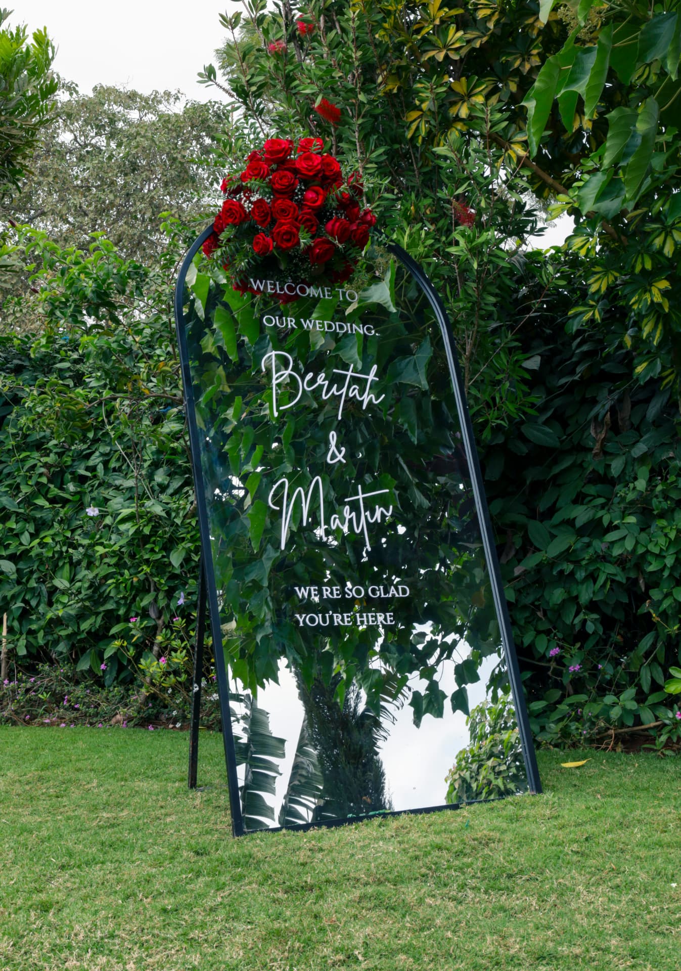 Mirror wedding welcome sign topped with red roses in the garden