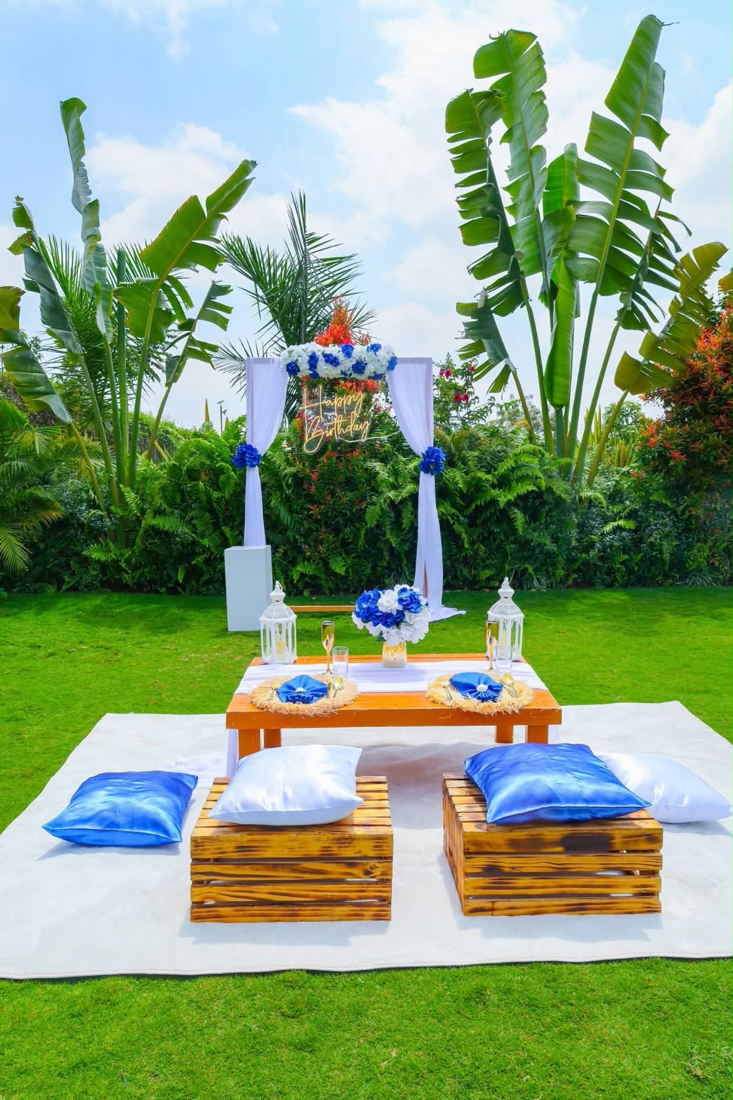 Clear blue and white birthday picnic setup with cushions, lanterns and flowers on the sunny garden lawn