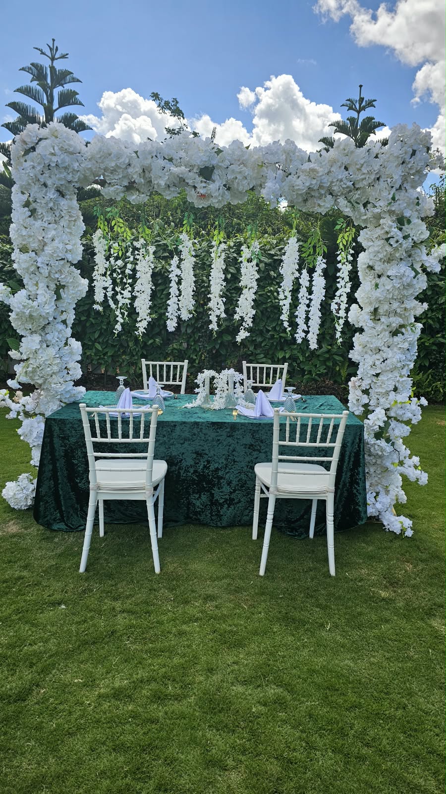 White floral canopy over an emerald dressed table with white Chiavari chairs on the garden lawn